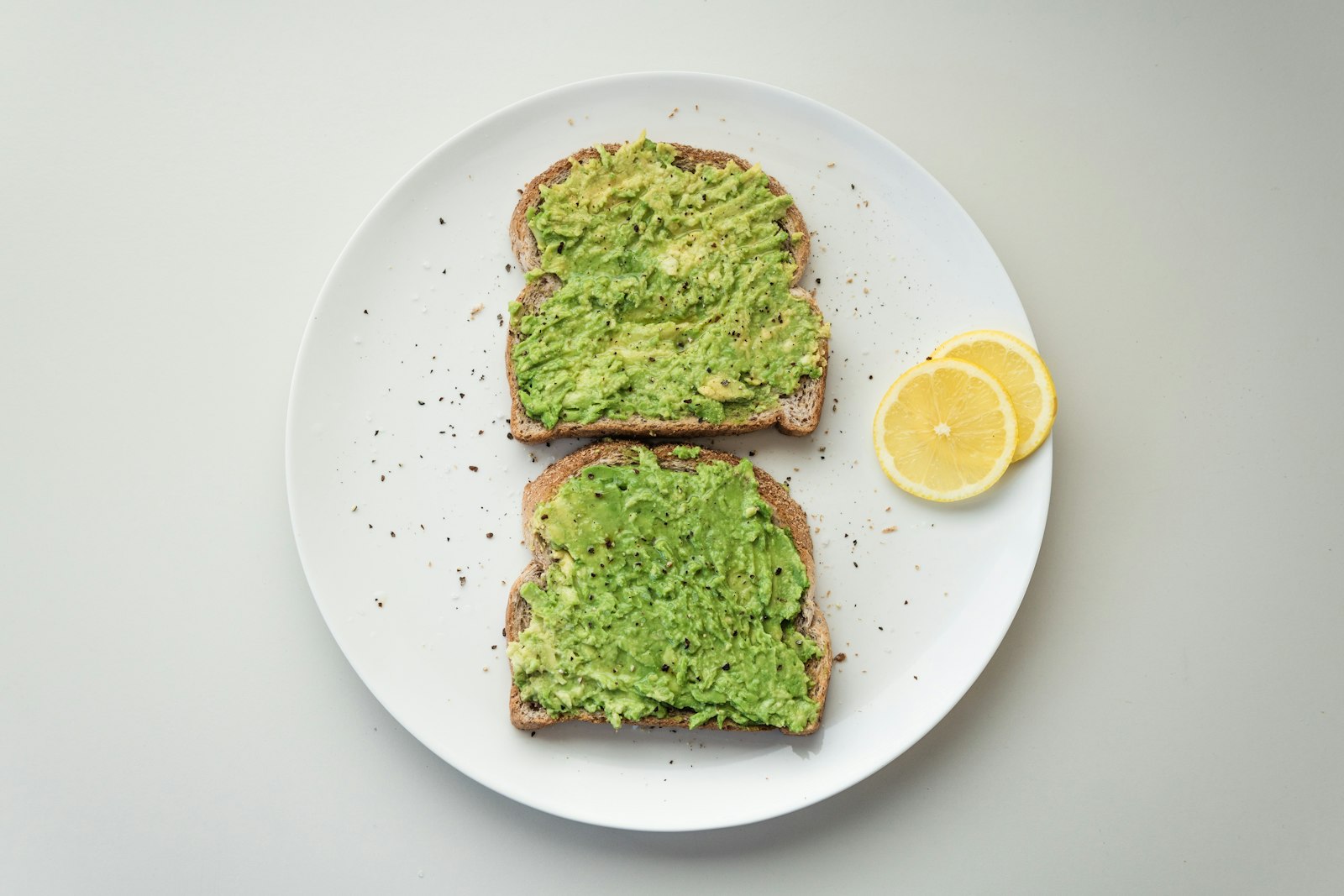 Two slices of toast topped with plain mashed avocado on a white plate, with lemon slices