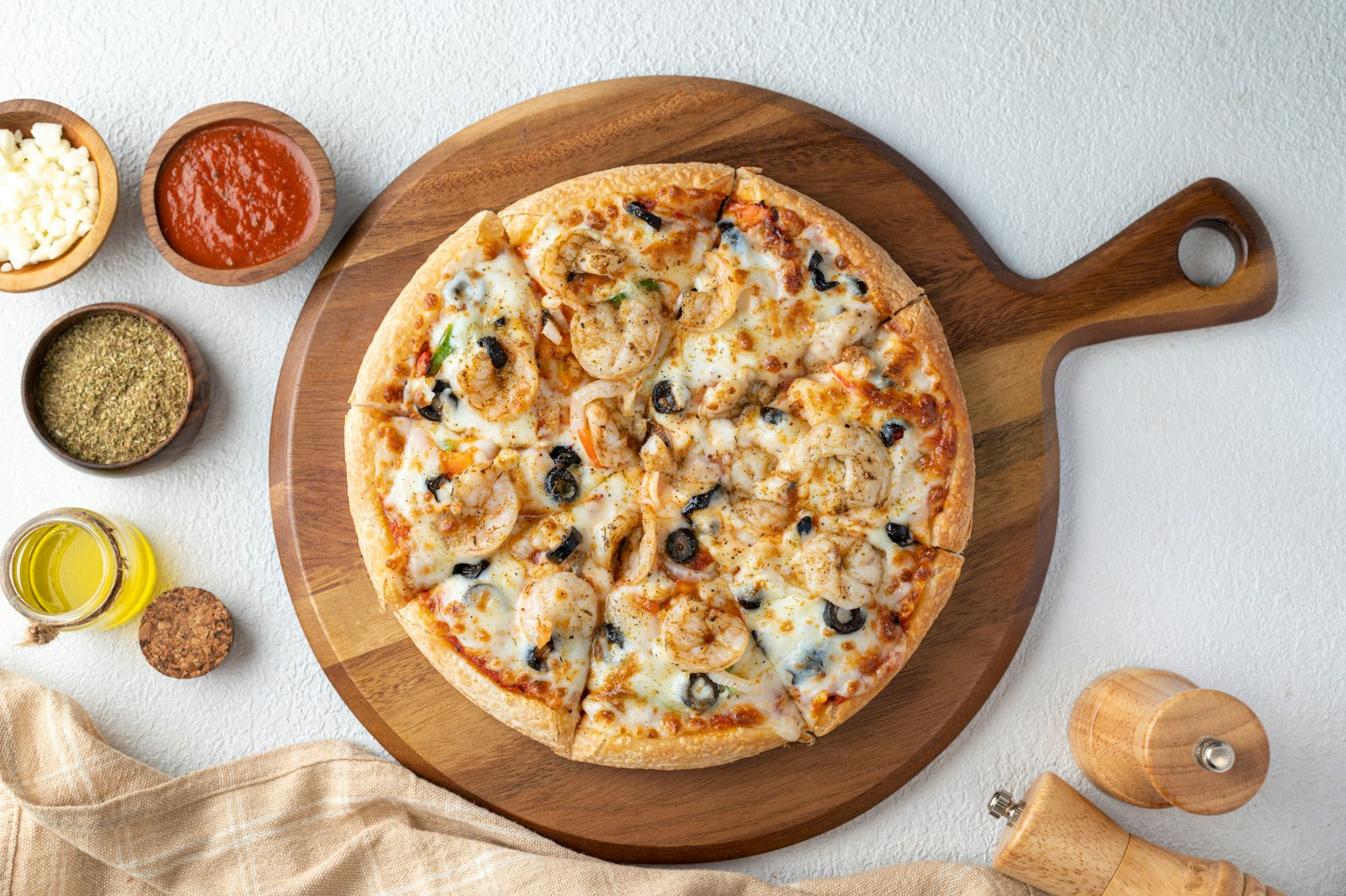 A pizza cut into slices on a round wooden board, with small bowls of oil, sauce and herbs beside it